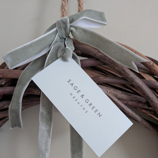 A close-up of a natural willow wreath base with a sage green velvet ribbon bow and a hanging sage green tag that reads 'SAGE & GREEN WREATHS'.