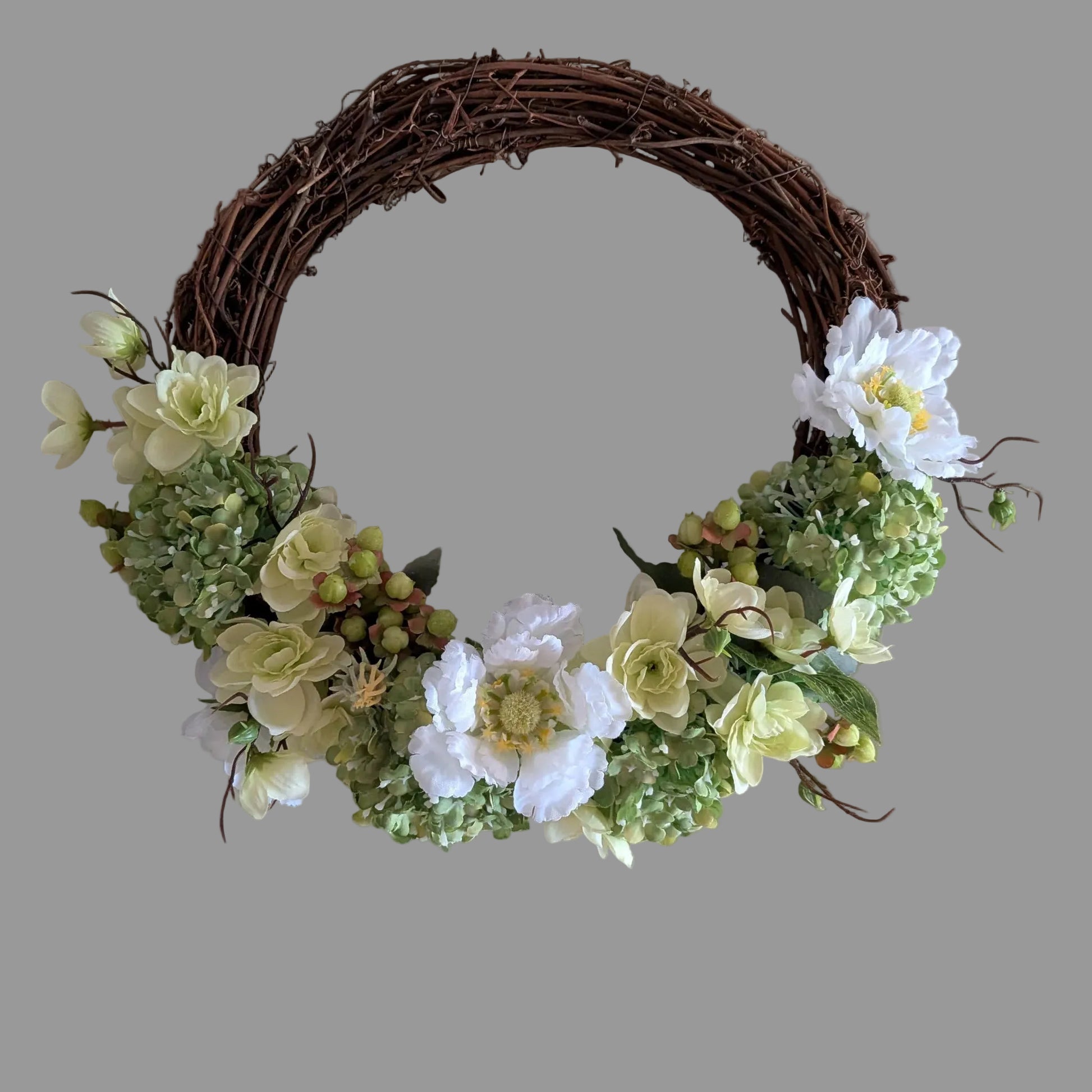 A rustic 30cm grapevine wreath decorated with white and pale yellow artificial flowers and green Vibernum -style blooms, shown against a plain gray background.