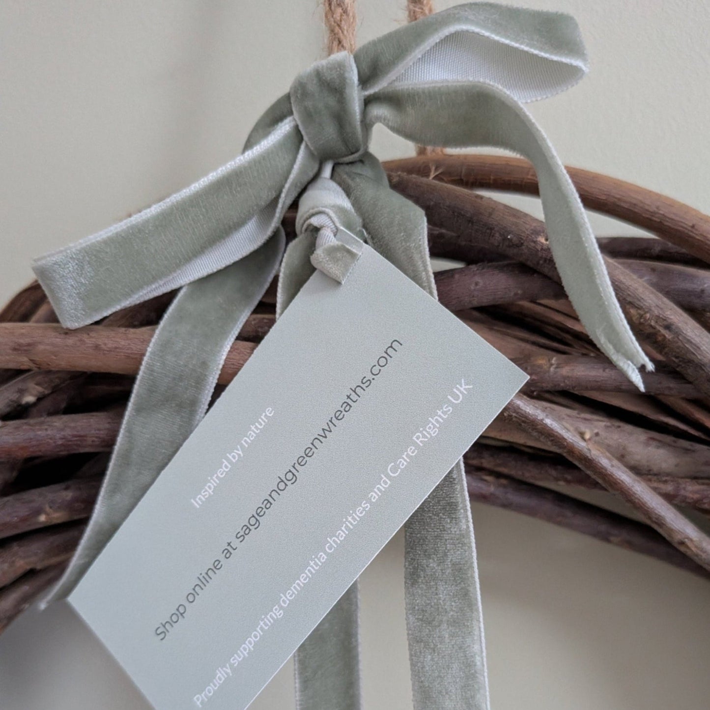 A close-up of a natural wicker wreath base decorated with a sage green velvet ribbon bow and a hanging tag that reads 'Inspired by nature' and 'sageandgreenwreaths.com'.